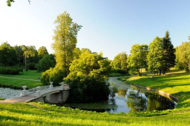 Summer day in Pavlovsky park, Pavlovsk, St. Petersburg, Russia. Beautiful views of the park, green trees, soft sun, flowing river, white bridge over the water. Nice evening in the park.