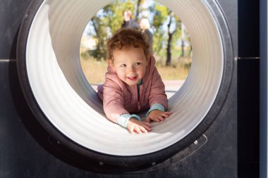 Küçük çocuk oyun parkındaki tünelden sürünerek geçiyor. İnsanlar