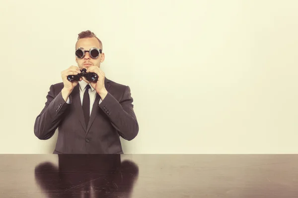 Businessman sitting at office desk with binoculars — Stock Photo, Image