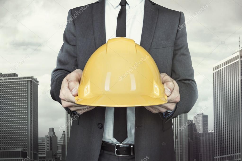 Businessman holding yellow helmet in hands on cityscape background