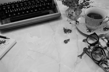 Eski moda bir daktilonun siyah-beyaz görüntüsü. Masanın üzerinde bir daktilo, bardak, not defteri, gözlük ve fotokopi alanı olan çiçekler..
