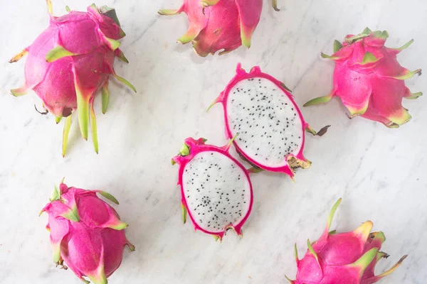 Top down view of bright pink dragon fruit or pitaya with one cut open to show the seeded flesh scattered on a textured white marble background.