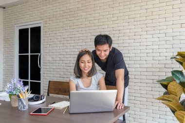 Genç Asyalı çift, modern mutfaktaki büyük ahşap masada otururken ve evde bilgisayar laptopuyla çalışırken rahat kıyafetler giyerler. Aşk, mutluluk, ev konseptinde çalışmak