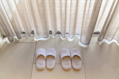 Beyaz perdeler ve yumuşak güneş ışığı ile beyaz terliklerin üst görünümü, tatil kavramı