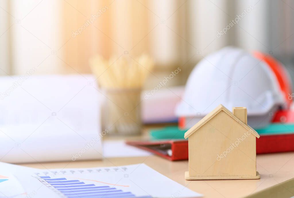 small wooden house or home with engineering tools, blueprint and white safety helmet on messy table, real estate, property and construction project concept