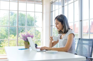 Gülümseyen genç Asyalı kadın günlük giysiler giyer bilgisayarla çalışır ve evde kahve fincanı tutardı. Ev konseptinde çalış