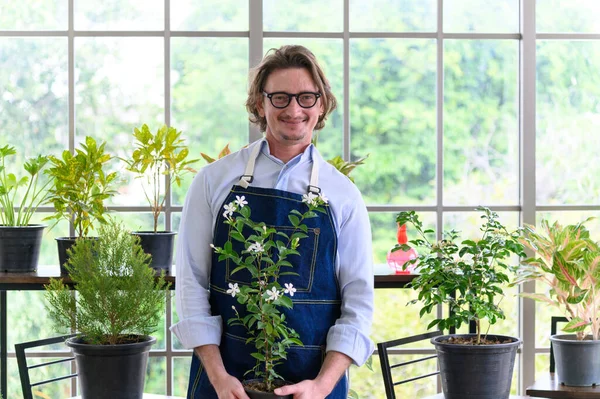 Portrait of happy gardener senior man wearing glasses holding small tree in plant pot as a hobby of home gardening at home