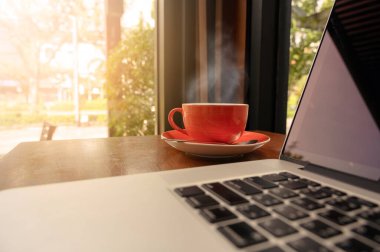 Kafe içindeki ahşap masada modern dizüstü bilgisayarlı bir fincan sıcak cappuccino kahvesi seçmeli olarak kahve fincanına odaklanmış. İş yeri, modern yaşam tarzı konsepti