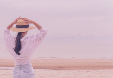 Açık havada, tropik plajda denize bakan hasır şapkalı genç Asyalı kadının yaz portresi, özgürlük zamanının, temiz havanın ve tatil tatilinin keyfini çıkar.