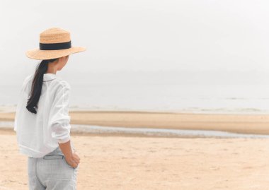 Açık havada, tropik plajda denize bakan hasır şapkalı genç Asyalı kadının yaz portresi, özgürlük zamanının, temiz havanın ve tatil tatilinin keyfini çıkar.