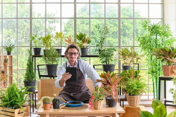 Happy senior man gardening wearing glasses and denim apron with digital tablet sitting in home gardening for looking who buys the tree. Small business concept.