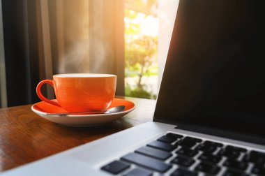 Kafe içindeki ahşap masada modern dizüstü bilgisayarlı bir fincan sıcak cappuccino kahvesi seçmeli olarak kahve fincanına odaklanmış. İş yeri, modern yaşam tarzı konsepti