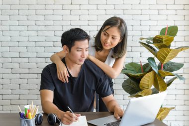 Genç Asyalı çift, modern mutfaktaki büyük ahşap masada otururken ve evde bilgisayar laptopuyla çalışırken rahat kıyafetler giyerler. Aşk, mutluluk, ev konseptinde çalışmak