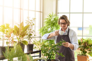 Bahçıvanlık yapan, gözlüklü ve kot önlüklü mutlu bir adamın portresi. Yaz sabahını elinde sıcak kahve tutarak evde bahçenin tadını çıkararak geçiriyor.