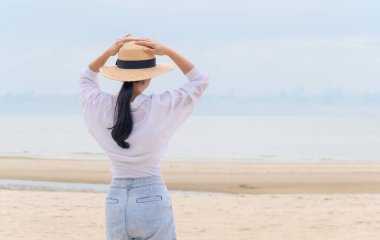 Açık havada, tropik plajda denize bakan hasır şapkalı genç Asyalı kadının yaz portresi, özgürlük zamanının, temiz havanın ve tatil tatilinin keyfini çıkar.