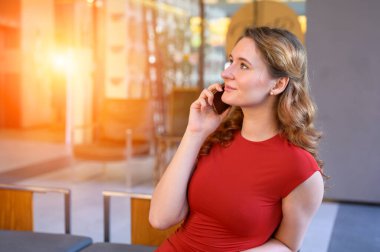 Modern bir lobide oturan zarif genç bir kadın cep telefonuyla konuşuyor. İş iletişimi, yaşam tarzı, teknoloji, bağlantı, modern çalışma hayatı kavramı