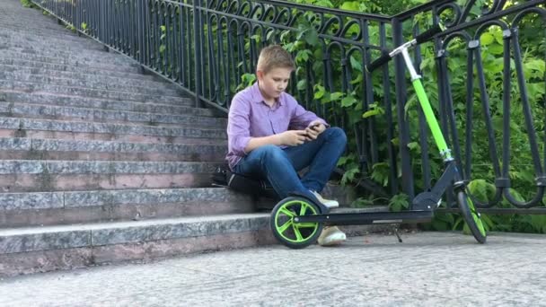 Мальчик сидит на лестнице в парке и пишет по телефону — стоковое видео