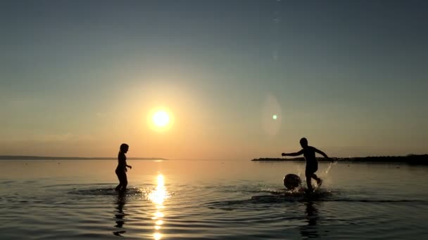 Barn spelar bollen i vattnet vid solnedgången. — Stockvideo