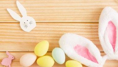 Mutlu Paskalya 'nın sembolü. Beyaz tavşan kulakları, pastel renkli paskalya yumurtaları ve açık tahta arka planda tahta tavşanlar. Metin için yer.