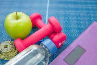 Two red dumbbells, an apple, a tape measure, a bottle of water and weight scales on a yoga mat. Concepts about fitness, sport and health