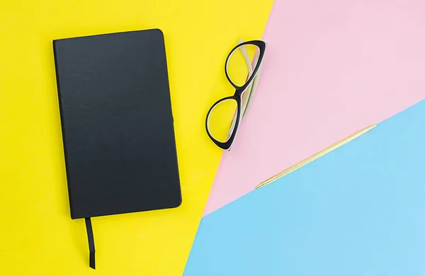 Caderno preto, caneta, óculos em fundo de papel amarelo, rosa e azul. Mesa de escritório de designer. Vista superior com espaço de cópia, flat lay. Contexto para o projeto — Fotografia de Stock