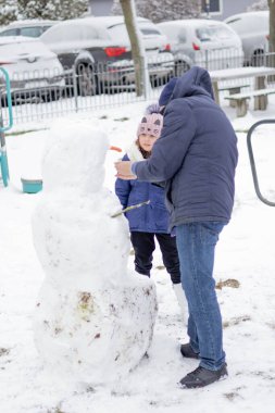 Bir çocuk ve bir yetişkin karlı bir parkta kardan adam yapıyorlar..