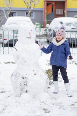 Kardan adamın yanında duran bir çocuk, karlı bir oyun parkında gülümsüyor..