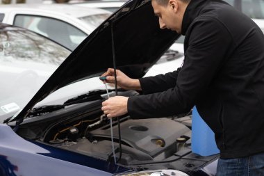 Bir adam diğer arabaların yanına park ederken kaputun altındaki silecek sıvısını kontrol ediyor..