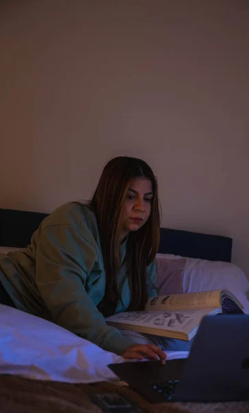 woman using a computer and reading a book lying on the bed