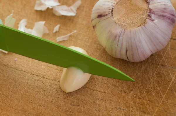 Peeled garlic segment being cut on a used cutting board with a green knife next to it there is a whole garlic head — Stock Photo, Image