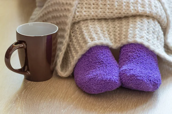 Cold concept with teacup near the legs which are covered with fluffy warm blanket and wearing fluffy warm purple socks — Stock Photo, Image