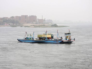 Nil, Mısır - 28 Mart 2015. Bir Mısır feribotu sabahın erken saatlerinde Nil 'e varıyor. Feribotta iki kamyon, araba, katır ve bazı insanlar var. Uzakta bir Mısır kasabasının belirsiz ana hatları var. 