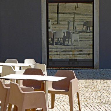 Modern sandalyeler, terasın masaları ve arkadaki dev pencerenin yansıması. 