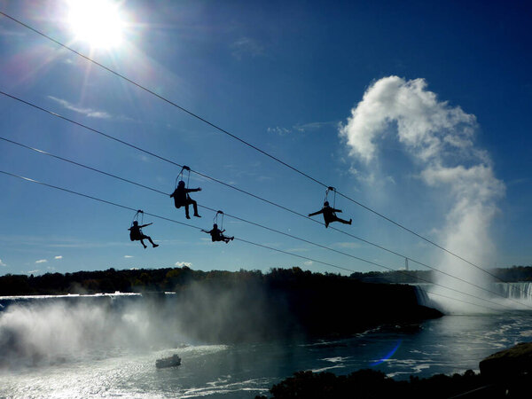Niagara Falls, Ontario, Canada - October 19, 2016: Four adventurous visitors ride a zip-line towards the Horseshoe Falls on a beautiful day in October. The mist at the falls builds a cloud in the distance