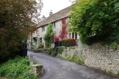 Cotswolds street in Castle Combe with charming stone cottages, featuring rustic architecture and steep, sloped roofs. The facade is adorned with climbing plants and vibrant red five-leaved ivy. 