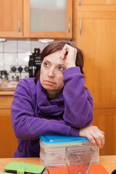 Middle-aging woman is sitting undecided in the kitchen.