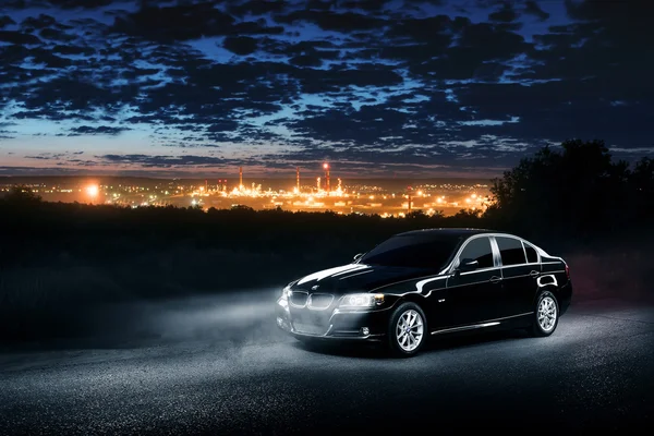 BMW E90, svart bil, står i den mørke tåkeskogen med utsikt over byen om natten. – stockfoto