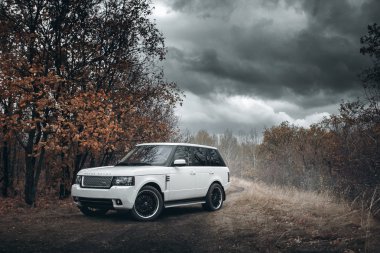 Beyaz araba Land Rover Range Rover gündüz dramatik bulutlarda şehir dışında duruyor.