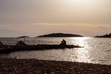 Gün batımında taş iskelede oturmuş, Hırvatistan 'ın Rogoznica kentindeki plajda deniz ve uzak adaları seyreden bir adam silueti.