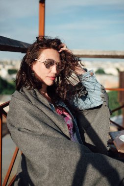 Orta yaşlı güzel bir kadın elinde bir fincan kahveyle çatıdaki bir kafede oturuyor..