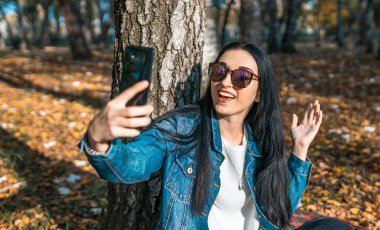 Sarı bir sonbahar parkında cep telefonuyla selfie çeken çekici genç bir kadın. Sevimli bir kız güneşli bir sonbahar gününde dışarıda yürürken kendi fotoğraflarını çekiyor. Günlük konsepti