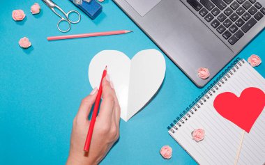 Bir kadının eli, kalp şeklinde bir karta yazar. Not defteri, defter, kalem, makas, mavi arka planda bir sürü kırmızı kalp. Sevgililer Günü tebrik kartı konsepti. Düz konum, üst görünüm, kopyalama alanı