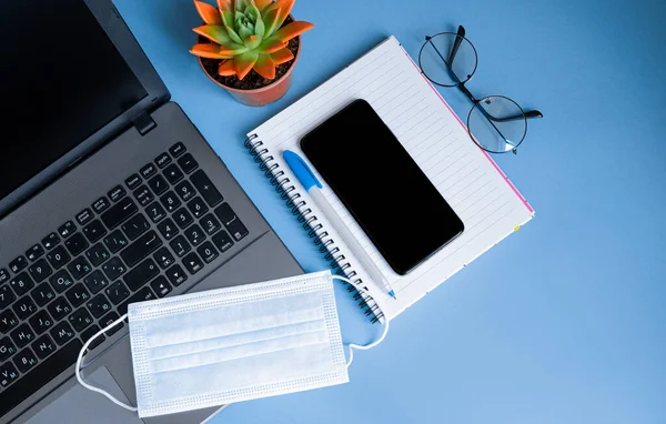 Blue desktop with computer, glasses, medical mask, smartphone, notebook and supplies.The concept of working in a home office. Top view with space for your text or app. Flat lay