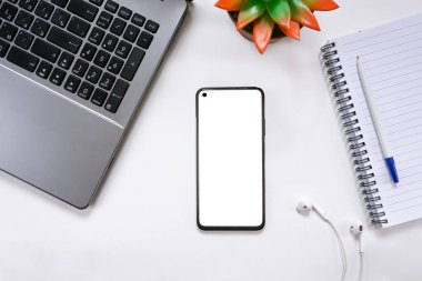 Beyaz ekran, dizüstü bilgisayar, kulaklık, not defteri ve manzara çiçeğiyle donatılmış akıllı telefonlu modern beyaz ofis masaüstü. Uzaktan kumanda konsepti, serbest çalışma. Üst görünüm, alanı kopyala.