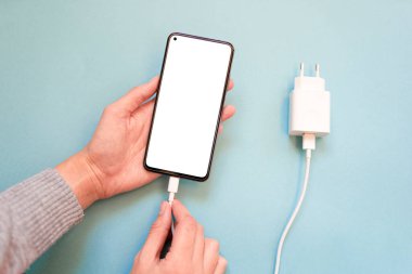 Akıllı telefonunuzu USB kablosu ve güç kaynağıyla mavi arka planda şarj etmek USB kablosunu, üst görüntüyü bağlar. Beyaz ekranlı akıllı telefonun silueti kabloyla sarkıyor. Kablolu şarj. kopyalama alanı. en üstteki görünüm