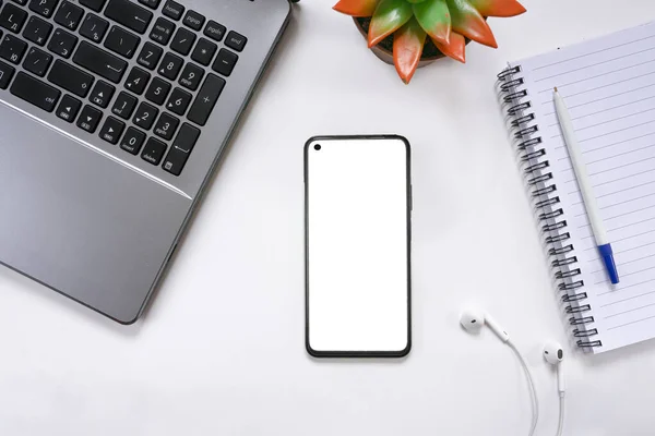 Beyaz ekran, dizüstü bilgisayar, kulaklık, not defteri ve manzara çiçeğiyle donatılmış akıllı telefonlu modern beyaz ofis masaüstü. Uzaktan kumanda konsepti, serbest çalışma. Üst görünüm, alanı kopyala.