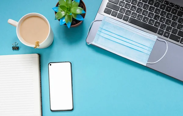Bilgisayarlı ofis masası, akıllı telefon, not defteri, kahve fincanı, tıbbi maske, kapalı mekan bitkileri, açık mavi arka planda mavi tıbbi maske. Karantina döneminde evde uzaktan çalışma kavramı. Boşluğu kopyala