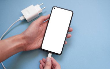 Bir akıllı telefonu USB kablosu ile şarj etmek ve bir güç ünitesini mavi arka planda bir USB kablosuna bağlamak, üst görünüm. Beyaz ekranlı akıllı telefonun silueti, USB kabloyla yüklenmiş..