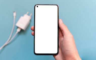 Elinde mavi arka planda beyaz ekran modeli olan akıllı telefon. Arka planda beyaz kablolu akıllı telefon şarjı var. kopyalama alanı. en üstteki görünüm