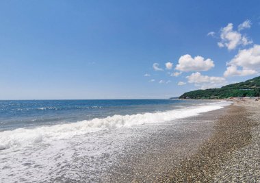 Deniz taşlı sahildeki dalgaların ve mavi gökyüzünün manzarası. Güneşli bir günde kayalık dağları ve berrak suları olan gök mavisi bir sahil. Rusya, Krasnodar Bölgesi